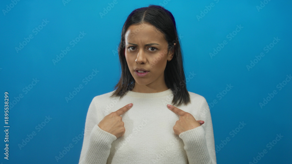 Obraz premium Woman pointing to herself with a questioning expression against a solid blue background, conveying curiosity and self-awareness, dressed in a white sweater.