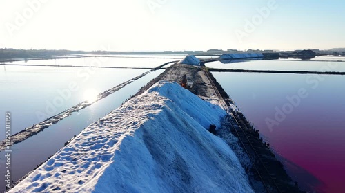 Perspective view of sea salt piles along narrow gauge railway tracks run alongside the salt heaps. Pink water ponds, bright sun reflecting on clear water.