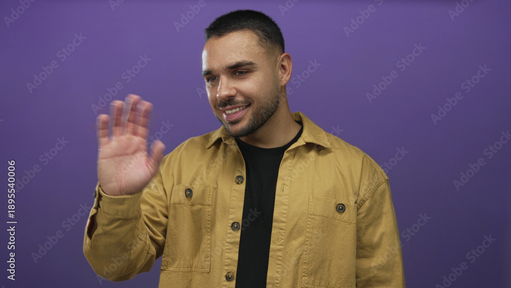 Fototapeta premium Man wearing mustard shirt and black tee with trimmed beard waving hand and smiling broadly in studio; joy.