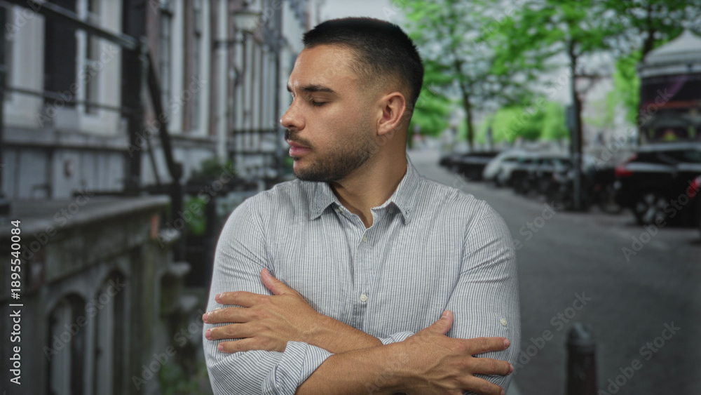 Fototapeta premium Young hispanic man in gray shirt crossing arms on city street lined with parked cars and leafy trees; rejection.