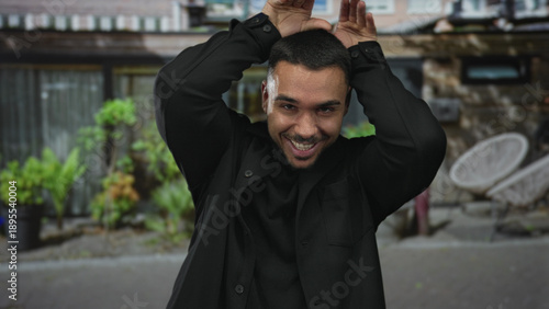 Hispanic man raises hands to head in street entrance of urban building while smiling broadly at camera; joy playfulness.