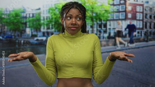 Photos African american woman in green turtleneck crop shirt with exposed midriff and braided hair smirks standing by a canal on an amsterdam street; skepticism