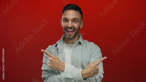 Quadro su tela Hispanic man with a joyful smile points in both directions against a vibrant red background, capturing an enthusiastic and engaging gesture in a studio setting