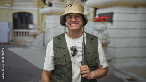 Quadro su tela Man holding fishing rod with bare hand, smiling in front of building, wearing fishing vest and bucket hat; leisure contentment