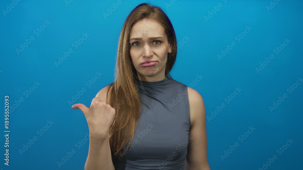 Fototapeta premium Woman in sleeveless gray top frowning with dramatic thumb pointing to side in blue studio setting; disgust.