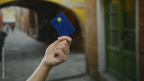 Obraz na plátně Hand of a caucasian man holding a blue credit card outdoors on a city street with green doors and archways in the background, conveying urban financial activity