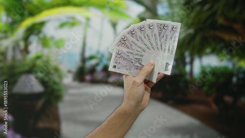 Photography Man's hand holding singapore dollars outdoors in a park setting, showcasing currency against a lush green backdrop, emphasizing finance and outdoor environment