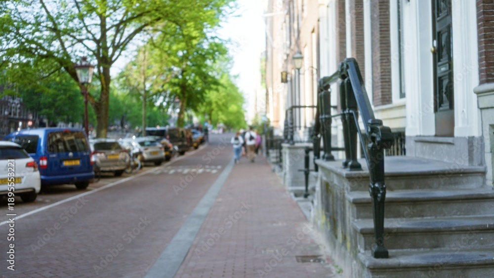 Fototapeta premium Two people walking along a blurred european street with cars parked beside and a bokeh effect on a sunny day, creating an urban and serene outdoor scene.