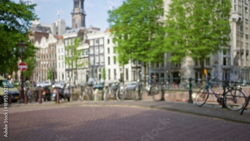 Bild auf Leinwand Blurred view of amsterdam's iconic canal-side architecture with bicycles lined up, trees, and cobblestone street creating a serene urban scene under sunny skies