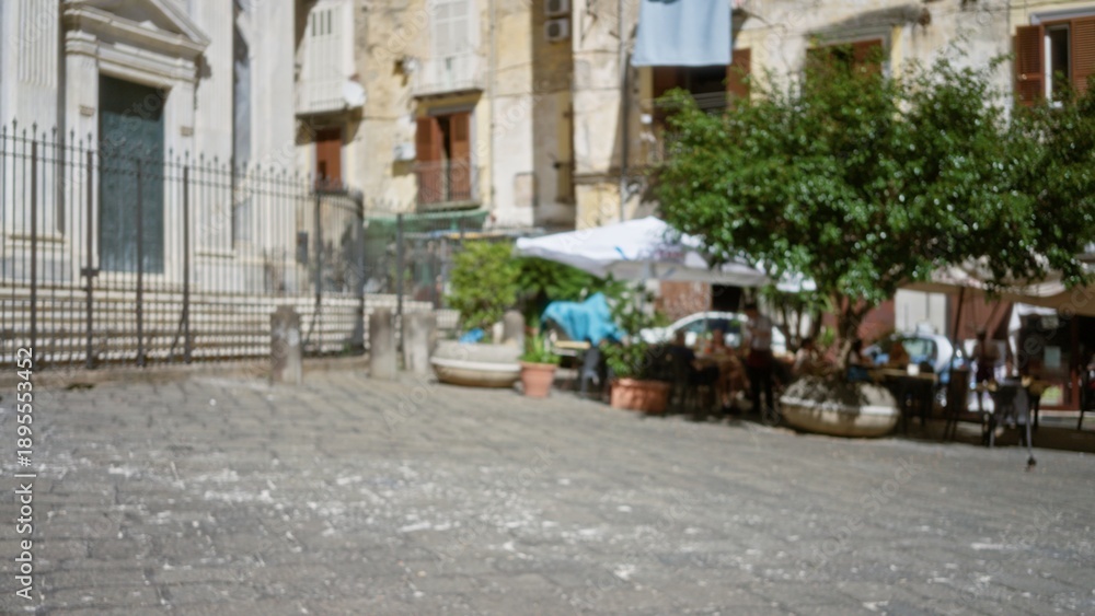 Obraz premium Defocused city plaza scene with cobblestone pavement, potted plants and cafe umbrellas in soft bokeh; background backdrop copyspace calm.