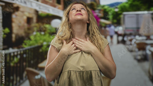 Φωτογραφία Blonde woman clasping hands over head on street terrace outside restaurant, head bowed then pressing hands to chest; relief
