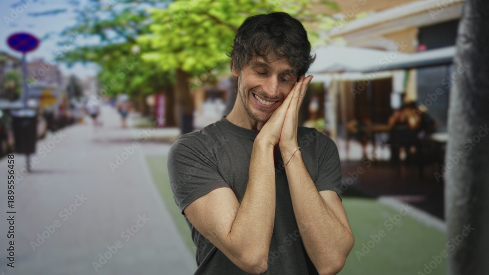 Fototapeta premium Young man wearing gray shirt clenching fists on street terrace of busy restaurant under green tree canopy; triumph.