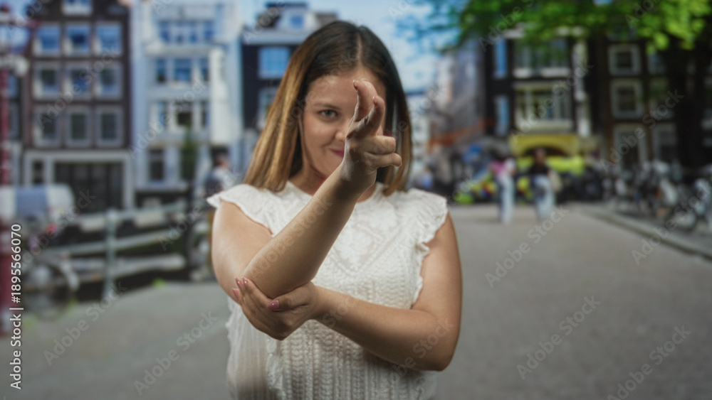Fototapeta premium Woman making finger to lips and a pointing gesture on street in front of canal houses with uncovered fingers and forearm visible; silence secrecy.