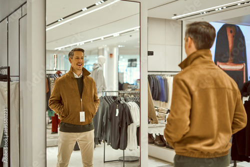 Fotografie Man Trying On Brown Suede Jacket in Fashion Mall Store, Smiling At His Reflectio