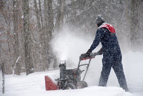 A side view of a man wearing winter coveralls using a snow blower to clear the driveway during a snowstorm

