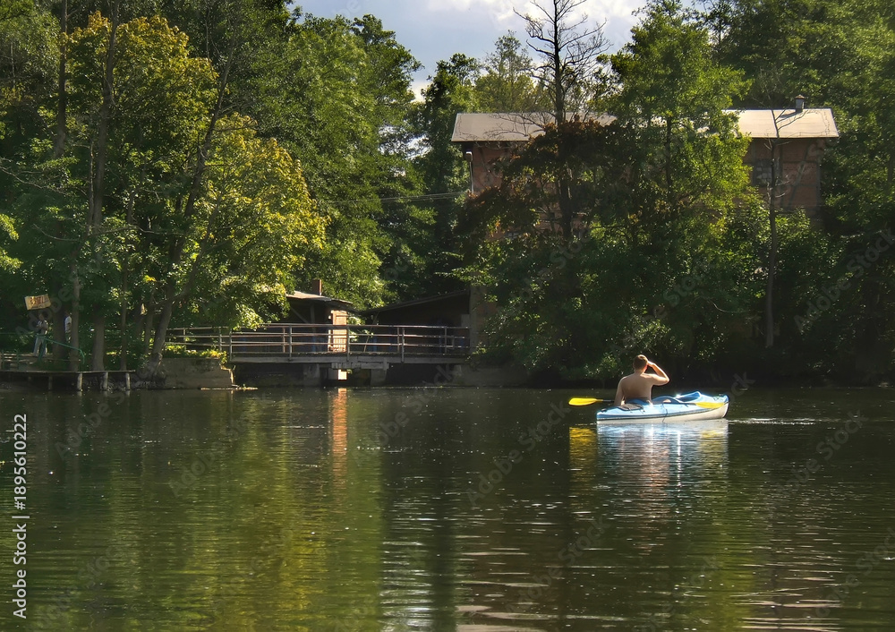 Obraz premium An unrecognizable person in a kayak seen from behind paddling on a beautiful and wild Krutynia river surrounded by dense green forest. Solitude and nature recreation in Poland.
