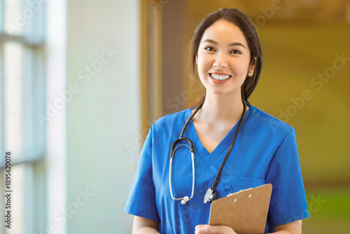 Bild auf Leinwand Asian female in blue scrubs standing in bright clinic corridor holding clipboard