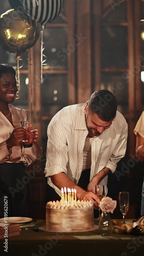 Happy young man celebrating birthday with friends, blowing out candles and making a wish before toasting with champagne