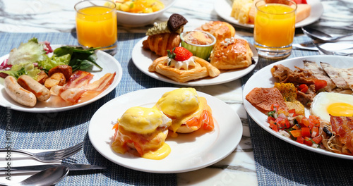 Gourmet breakfast spread featuring eggs benedict with hollandaise sauce, fresh orange juice, and assorted pastries on a marble table. Luxury hotel dining experience, healthy morning meal, and