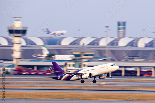 Passenger airplane takes off from busy airport runway, with terminal and control tower visible in background. scene captures dynamic motion of air travel