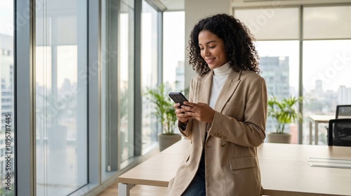 Young professional woman in beige blazer smiling while checking smartphone in bright modern office with city view and plants