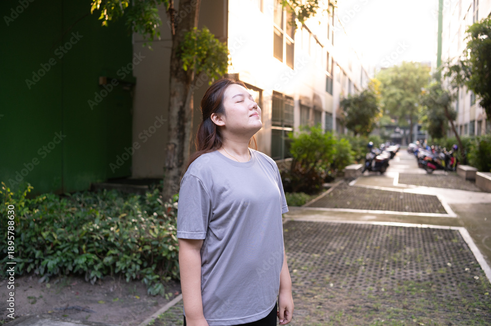 Fototapeta premium A person stands outdoors with eyes closed, basking in the sunlight on a leafy pathway beside parked motorcycles in a residential area.
