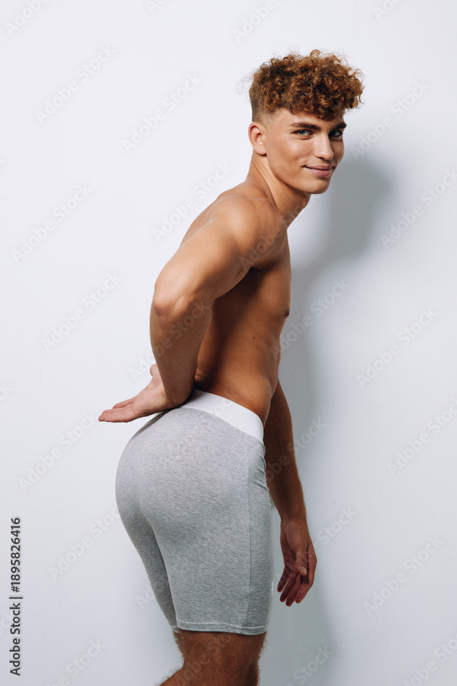 Fototapeta premium Young man with curly hair posing shirtless in gray shorts against white background, showing athletic build and confident expression in studio lighting.