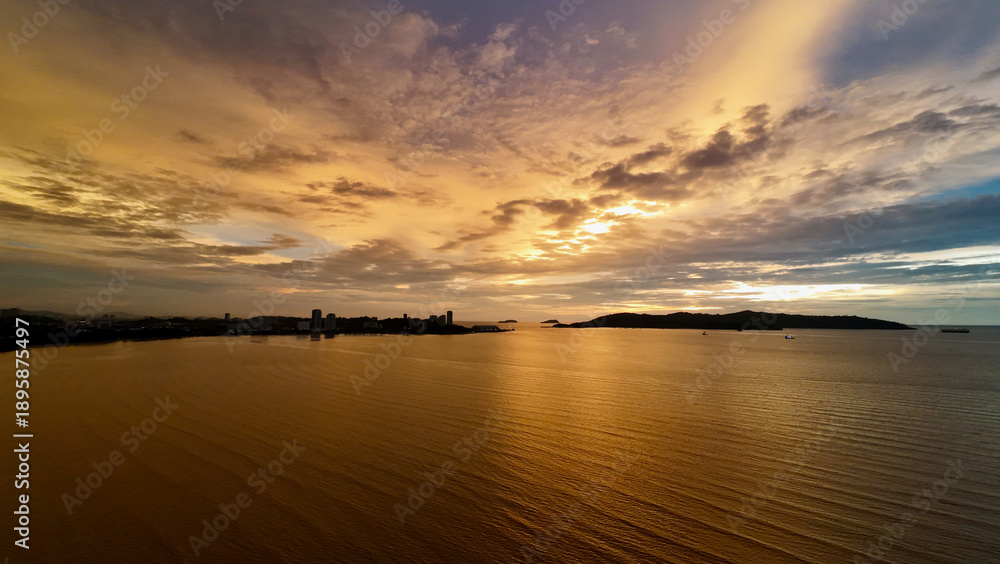 Obraz premium Golden sunset over tropical sea with city skyline on the horizon. Aerial view shows glowing water, dramatic clouds and urban coastline silhouetted against evening sky.