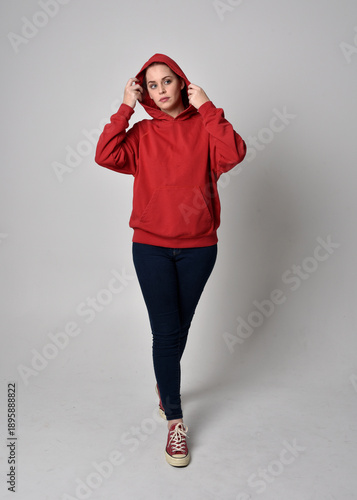 full length figure of female model wearing  basic contemporary casual clothing, red hoodie jumper, skinny jeans and sneakers. standing pose walking towards camera,  isolated on studio background.

