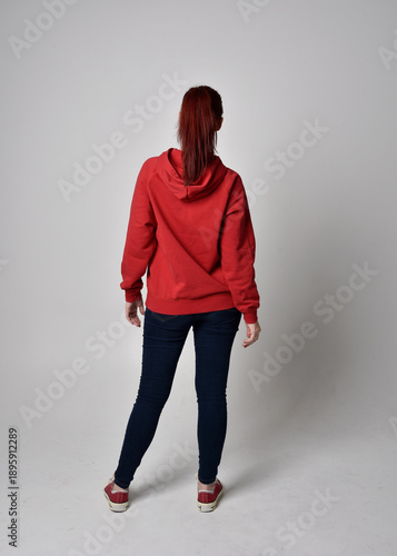 full length figure of female model wearing  basic contemporary casual clothing, red hoodie jumper, skinny jeans, sneakers. standing pose walking away back view,  isolated white studio background.
