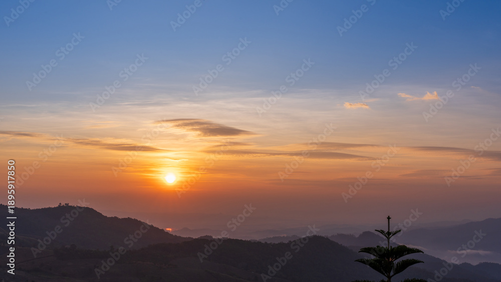 Fototapeta premium Sunrise over mountain layers with soft mist and tree silhouette creating a peaceful morning scene.