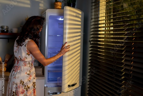 Evening kitchen activity, Lady opens fridge late at night, Woman in floral outfit seeks jar in darkness, Night example of woman retrieving item from cold storage
