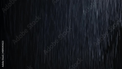 A dark, moody image depicting a heavy downpour of rain against a black background. Raindrops stream downward. The scene suggests a night scene