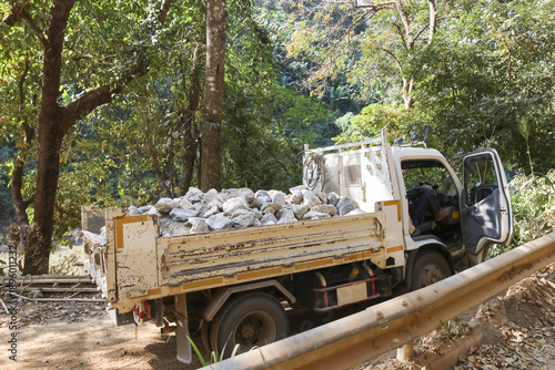 Rugged white truck hauls heavy cargo of stone on rural dirt road through dense forest. Transportation work showing construction and industry in remote area