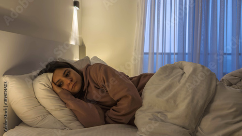 Young woman lying on bed at night with sad expression low mood dim bedroom light emotional exhaustion mental health struggle isolation loneliness depression concept fatigue insomnia