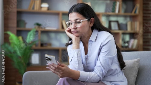 Tired bored young woman using smartphone while sitting on sofa at home. Exhausted sad female browsing social media, scrolling feed reading posts, wasting time on mobile phone in living room. Close up