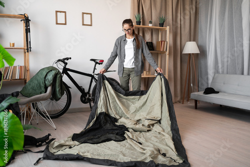 Canvas-taulu Young woman traveler packing camping tent while preparing for hiking trip at hom