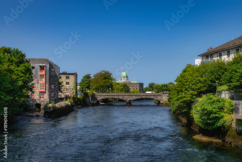 sunlit river with stone bridge, dome historic riverside scene framed by leafy trees and aged buildings, calm water reflecting clear sky, street photographer capturing architectural details,