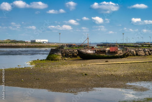 Low tide harbor stranded wooden trawler grounded on rocky mudflat, distant breakwater and calm sky, weathered paint, seaweed textures, quiet coastal solitude and nostalgic atmosphere