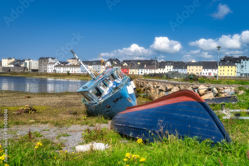beached blue and red boats on grassy shore with yellow wildflowers, tilted hulls with flaking paint, distant pastel town facades across calm estuary, sunlit summer day with scattered clouds,
