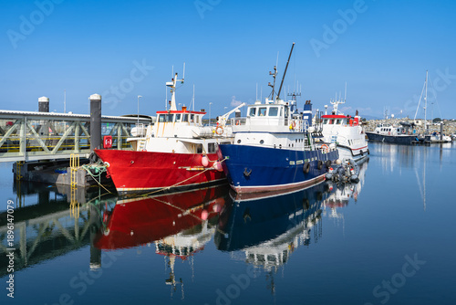 Peaceful waterfront with colorful trawlers and glossy reflections on calm surface in Galway, Rossaveel, Ireland. Idyllic harbor showcasing moored fishing boats beneath clear sky