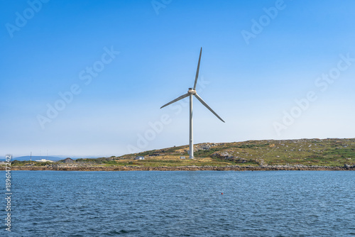 Wind turbine rising above coastal water near Rossaveel, Galway in Ireland. Rocky shoreline in midground, gentle waves in foreground, clear blue sky, symbol of renewable energy