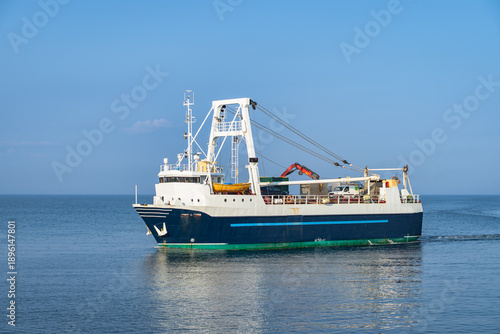 Working trawler underway on open sea near Inis Mor Island, Galway in Ireland. Deck cranes and winches poised for net deployment. Calm water with gentle ripples, distant coastline on horizon