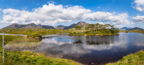 Panoramic mountain lake with Pine Island in Connemara, Galway, Ireland. Calm reflective water under dramatic clouds. Distant rocky peaks, grassy marsh foreground and scattered peat hummocks