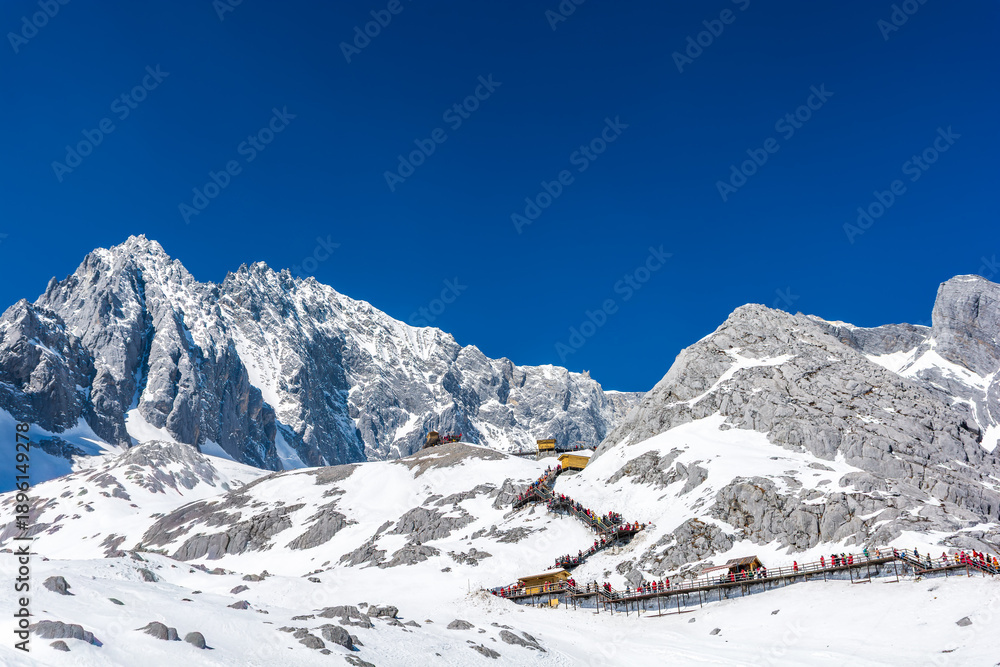 Naklejka premium Majestic view of Jade Dragon Snow Mountain with tourist walkway under a clear blue sky in Lijiang, Yunnan, China. Stunning winter alpine landscape and famous high altitude travel destination.