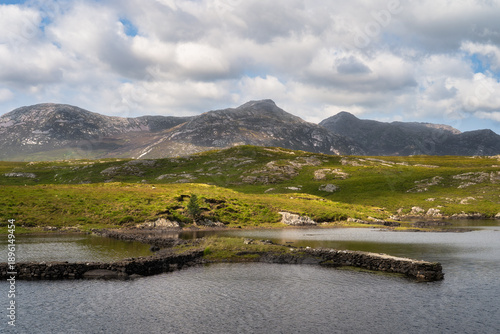 Rocky moorland with distant mountain ridge in Connemara, Galway, Ireland. Patchy grass and stone outcrops, marshy pools and dramatic clouds. Textured terrain conveying raw highland