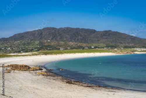 Curved Dogs Bay in Connemara, Galway, Ireland, with turquoise water and green hills. Small coastal village along shore, crescent beach with rocky outcrops. Clear shallow water and bright midday sky