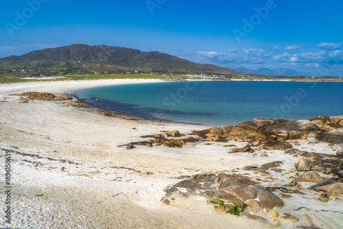 Rocky shoreline with clear turquoise sea and white sand of Dogs Bay in Connemara, Galway, Ireland. Scattered kelp beds and tidal pools. Rugged foreground leading to rolling hills