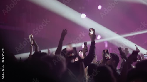 Video of a large crowd at a music festival or rock concert. Fans hold up smartphones to record the band on stage amid dramatic beams of light and smoke. A modern live event experience.