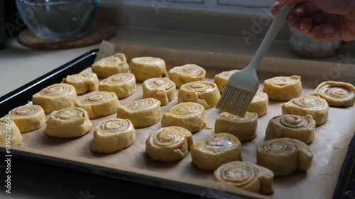 glazing raw unbaked cinnamon rolls on tray before put them in oven, baking at home, daylight, woman hand with food brush